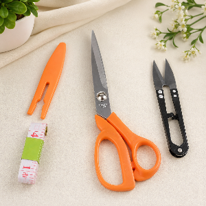 Orange scissors, black shears, and a measuring tape on a light surface with plants in the background.