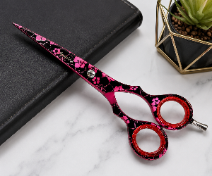 Pink and black cherry blossom patterned hair scissors resting on a black leather folio, set on a white marble surface next to a gold geometric planter.